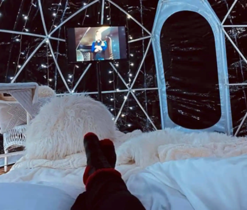 Cozy indoor scene with a person’s feet up, watching TV in a stylish igloo-like space with soft blankets and fairy lights.