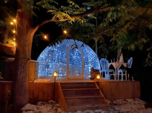 A cozy, illuminated geodesic dome surrounded by trees, with a table and chairs on a wooden deck.