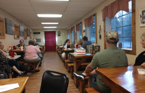 A cozy restaurant interior with wooden tables, patrons chatting, and a warm atmosphere. Windows with checkered curtains.
