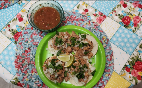Three tacos filled with shredded meat, onions, and cilantro, served with lime and a bowl of salsa on a colorful tablecloth.