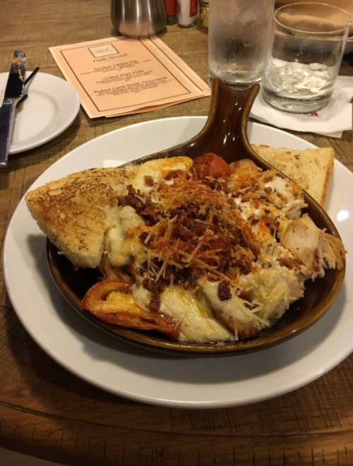 A baked dish with pasta, chicken, cheese, and bacon, served with toasted bread on a white plate.
