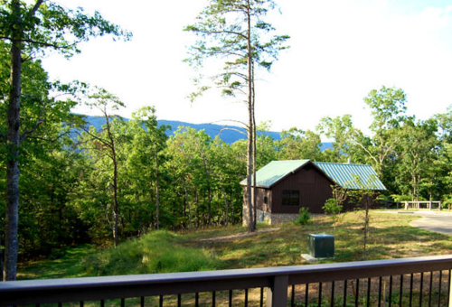 A cabin with a green roof surrounded by trees and mountains in the background on a sunny day.