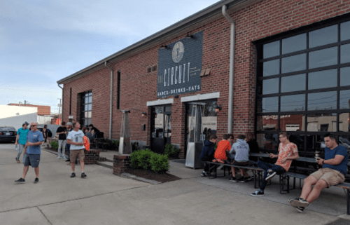 A brick building with a sign reading "Games, Drinks, Eats," featuring people outside and seated at benches.