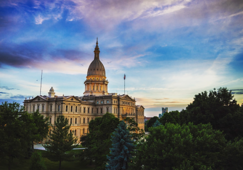 The Michigan State Capitol building at sunset, surrounded by trees and a colorful sky.