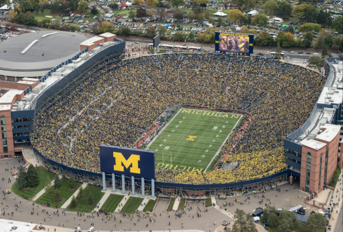 Aerial view of a packed Michigan Stadium filled with fans in maize and blue, with the field and scoreboard visible.