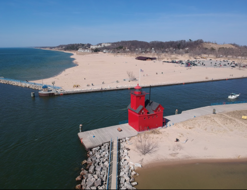 Aerial view of a red lighthouse on a pier, with a sandy beach and calm water in the background.
