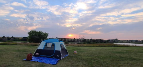 A tent set up on green grass with a sunset and clouds in the background, near a calm body of water.