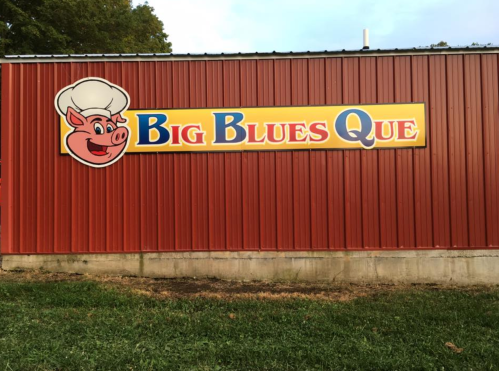Sign for "Big Blues Que" featuring a cartoon pig wearing a chef's hat, on a red metal building.