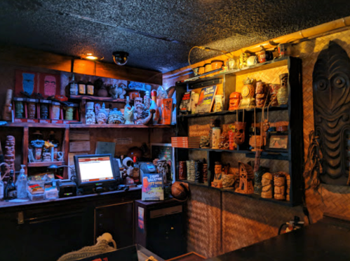 A dimly lit bar with tiki decor, shelves filled with colorful mugs, and a cash register on the counter.