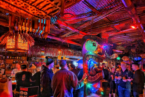 A vibrant tiki bar scene with colorful lights, a large tiki statue, and patrons enjoying drinks and conversation.
