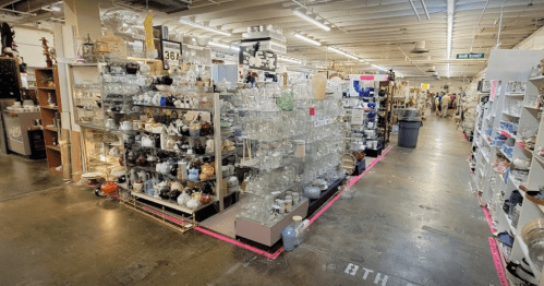A spacious thrift store aisle filled with shelves of glassware and various items, with shoppers in the background.