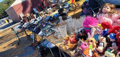 A vibrant flea market scene with tables displaying shoes, glassware, toys, and a doll, set against a rustic backdrop.