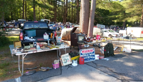 A bustling flea market scene with tables of various items for sale, parked cars, and trees in the background.