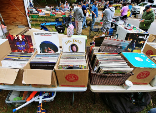 A collection of vinyl records displayed in boxes at a bustling outdoor market with people browsing in the background.