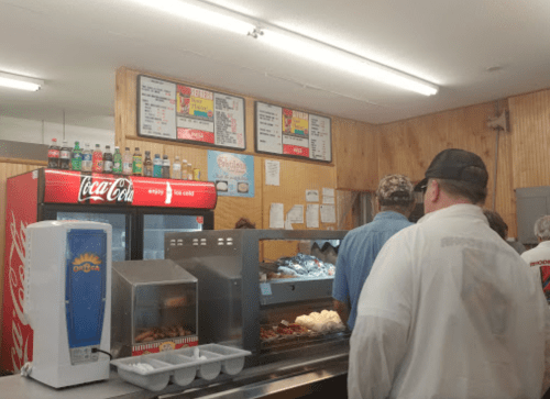 A busy diner with a Coca-Cola machine, menu boards, and customers waiting in line at the counter.