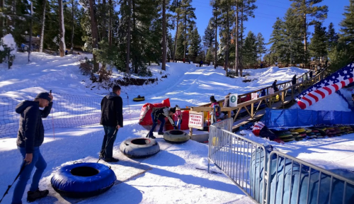 People preparing to go tubing on a snowy hill, with trees and a colorful ramp in the background.