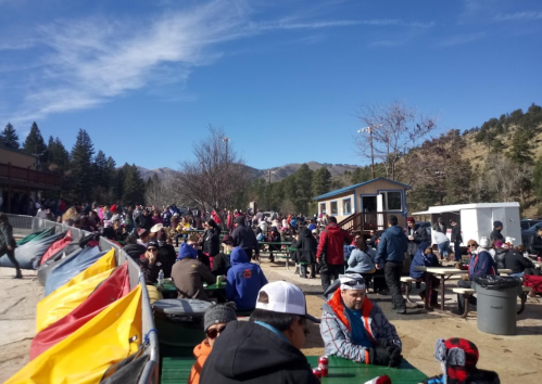 A crowded outdoor area with people sitting at picnic tables, surrounded by trees and mountains under a clear blue sky.