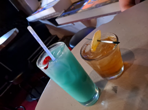 Two cocktails on a table: a blue drink with a cherry and a brown drink with a lemon slice and a straw.