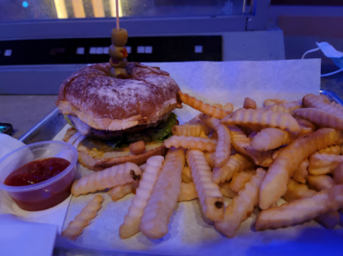 A burger with a skewer on top, served with a side of crinkle-cut fries and a small cup of ketchup.