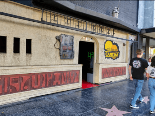 A group of people walks past a themed cantina with a decorative facade and a bright sign.