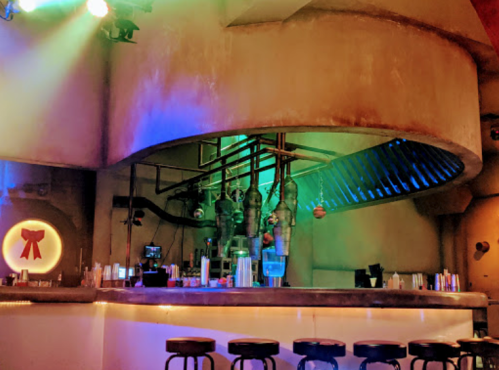 A modern bar with a curved design, colorful lighting, and industrial decor, featuring stools and a well-stocked counter.