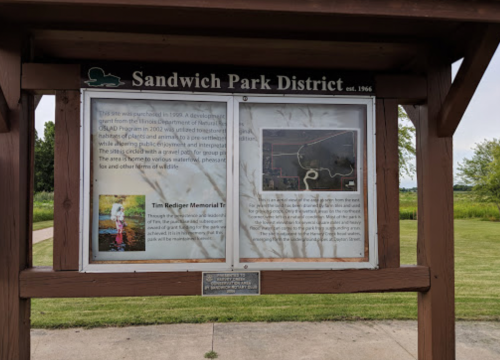 Sign at Sandwich Park District featuring information about the park and the Tim Rediger Memorial Trail. Green grass in background.