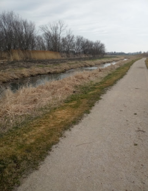 A gravel path alongside a small stream, with bare trees and dry grass on either side under a cloudy sky.