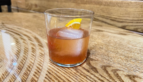 A glass of cocktail with a slice of orange and ice, placed on a wooden table.