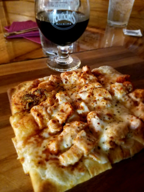 A wooden board with a cheesy pizza topped with chicken and spices, alongside a glass of dark beer.