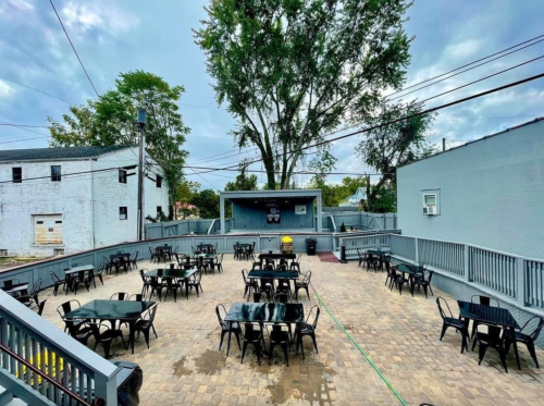 Outdoor patio with empty tables and chairs, surrounded by trees and buildings, under a cloudy sky.