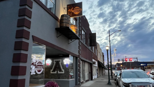 A street view of a building with a barrel sign, featuring a restaurant and storefronts under a cloudy sky.