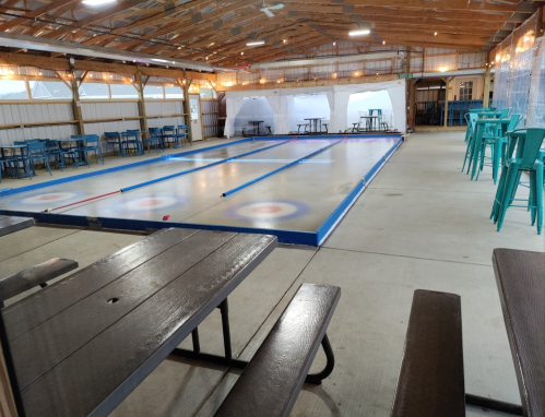 Indoor curling rink with blue lines and target circles, surrounded by tables and chairs in a rustic setting.