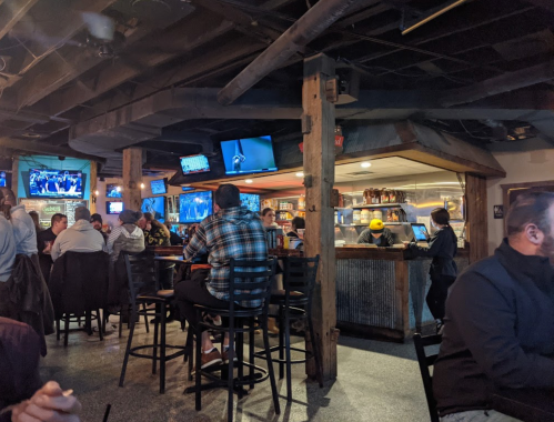 A lively bar scene with patrons seated at tables and a counter, multiple TVs showing sports in a cozy atmosphere.