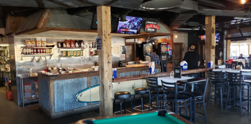 Interior of a casual bar with a wooden counter, dartboard, and pool table, featuring a variety of condiments and drinks.