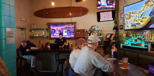 A bar scene with patrons at the counter, a bartender, and multiple TVs displaying sports and information.