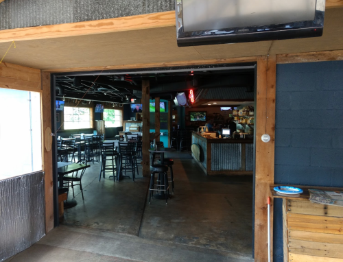 View of a rustic bar interior with empty tables and a counter, seen through an open doorway.