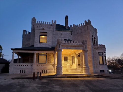 A historic stone building, Ashlar Hall, illuminated at dusk with a castle-like design and surrounding trees.