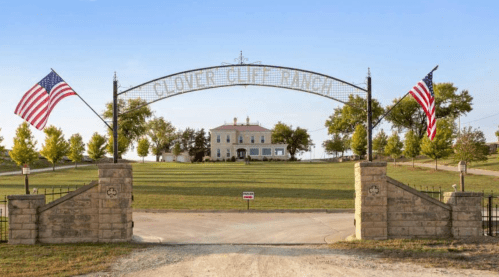 Entrance to Clover Cliff Ranch, featuring a grand gate and American flags, with a large house in the background.