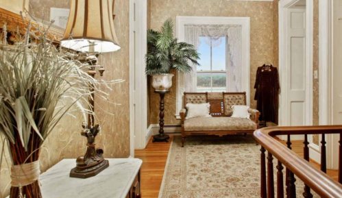 A vintage hallway with a lamp, decorative plant, and a wooden bench by a window, featuring ornate wallpaper and a rug.