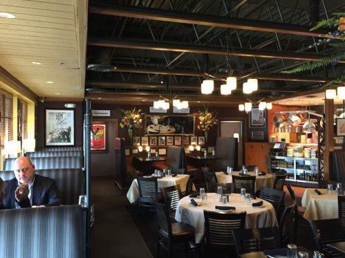 A cozy restaurant interior with tables set for dining, featuring artwork and decorative plants, and a man seated in the foreground.