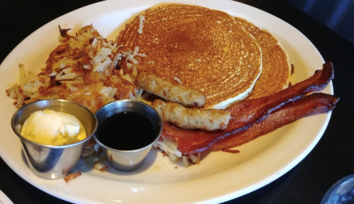 A breakfast plate featuring pancakes, hash browns, sausage links, bacon, and small cups of butter and syrup.