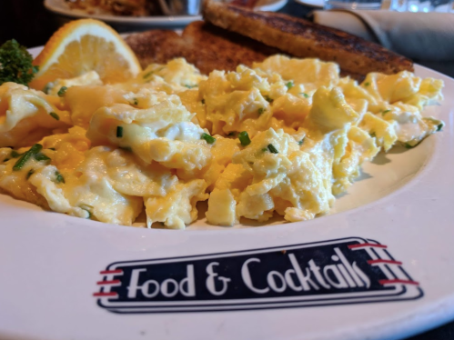 A close-up of a plate with scrambled eggs, garnished with chives and a slice of lemon, next to toast.