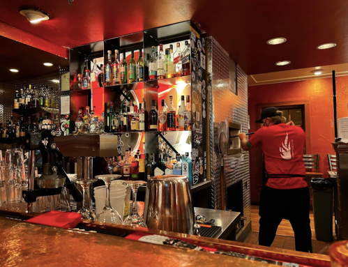 A bartender in a red shirt prepares drinks at a vibrant bar filled with colorful bottles and glasses.