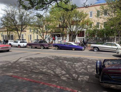A row of classic cars parked along a tree-lined street with people gathered nearby.