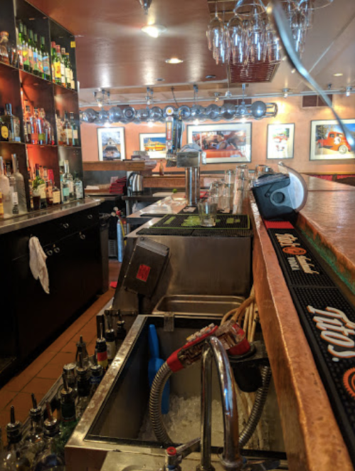 A bar area with shelves of liquor, a stainless steel sink, and a countertop with glasses and drink dispensers.