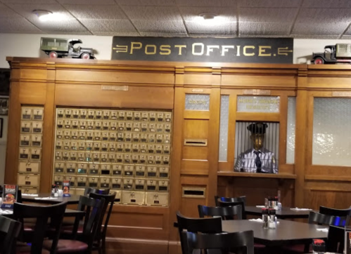 A vintage-style restaurant interior featuring a post office display with mailboxes and a server behind a counter.