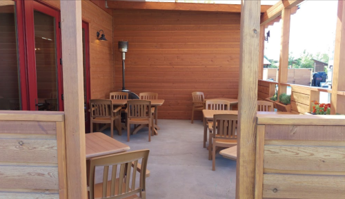 A wooden patio with several tables and chairs, surrounded by wooden walls and a sunny outdoor setting.