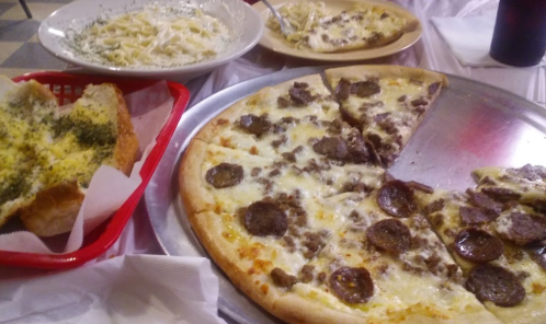 A table with a large pizza topped with sausage, garlic bread, and a bowl of creamy pasta.