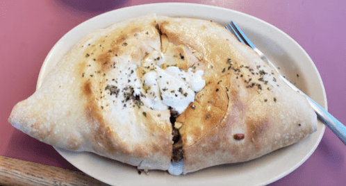 A large calzone filled with cheese and meat, topped with herbs, served on a plate with a fork beside it.