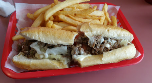 A red tray holds two cheesesteak sandwiches with fries, featuring beef, cheese, and toppings.